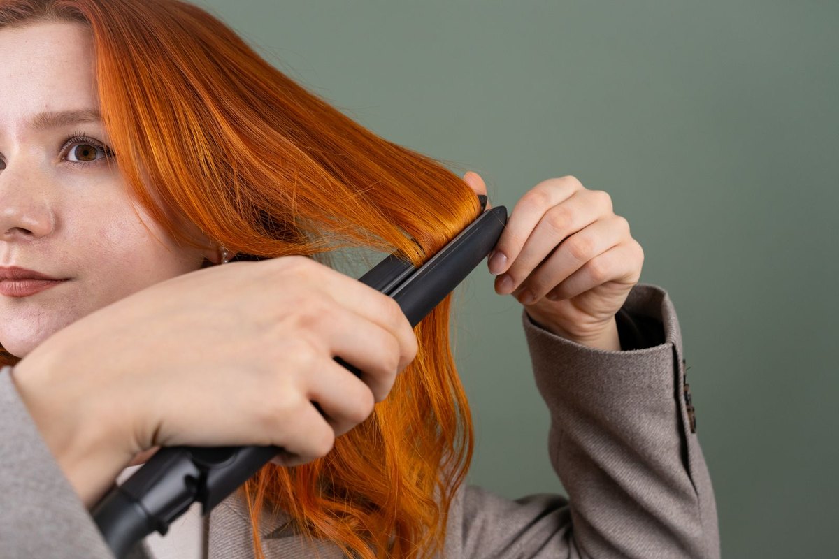 Close-up of a woman passing a black flat iron through a section of her long red hair