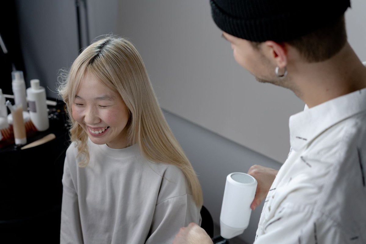 A hairstylist blow-drying a smiling woman’s hair with a white hair dryer in a salon